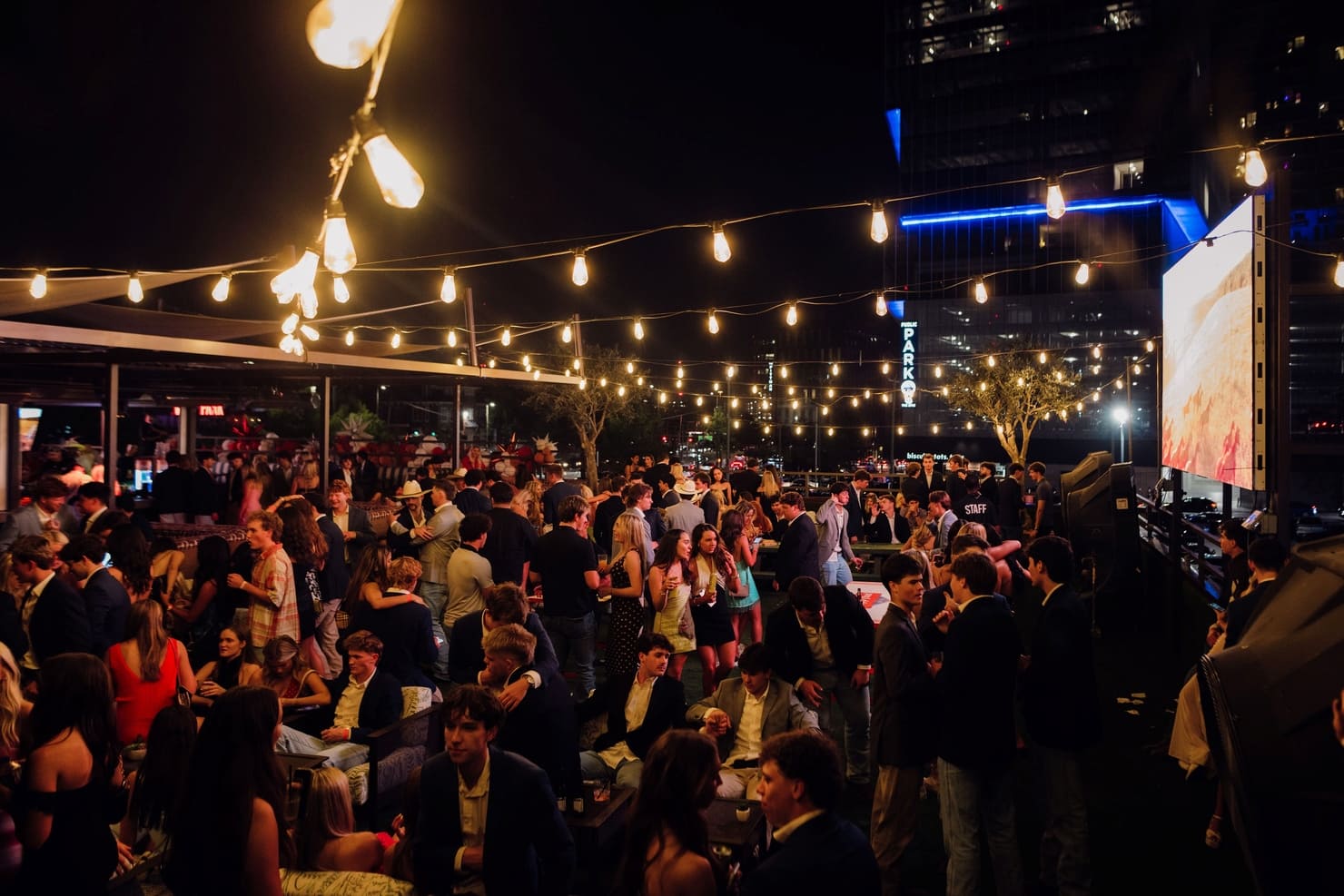 Rooftop nightlife party with large crowd under string lights in downtown city setting