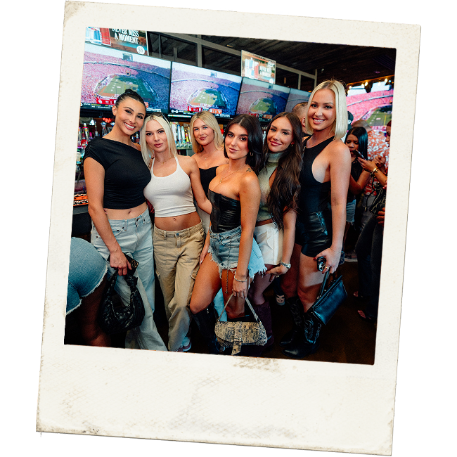 Group of six women posing at a sports bar with football games playing on multiple TV screens behind them.