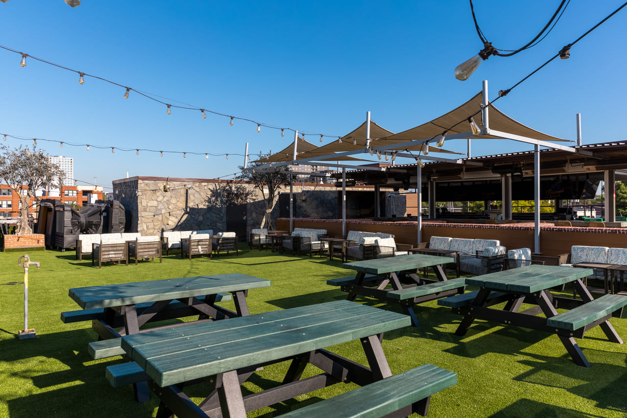 Outdoor seating area with green picnic tables and cushioned lounge chairs beneath tan shade sails and string lights.