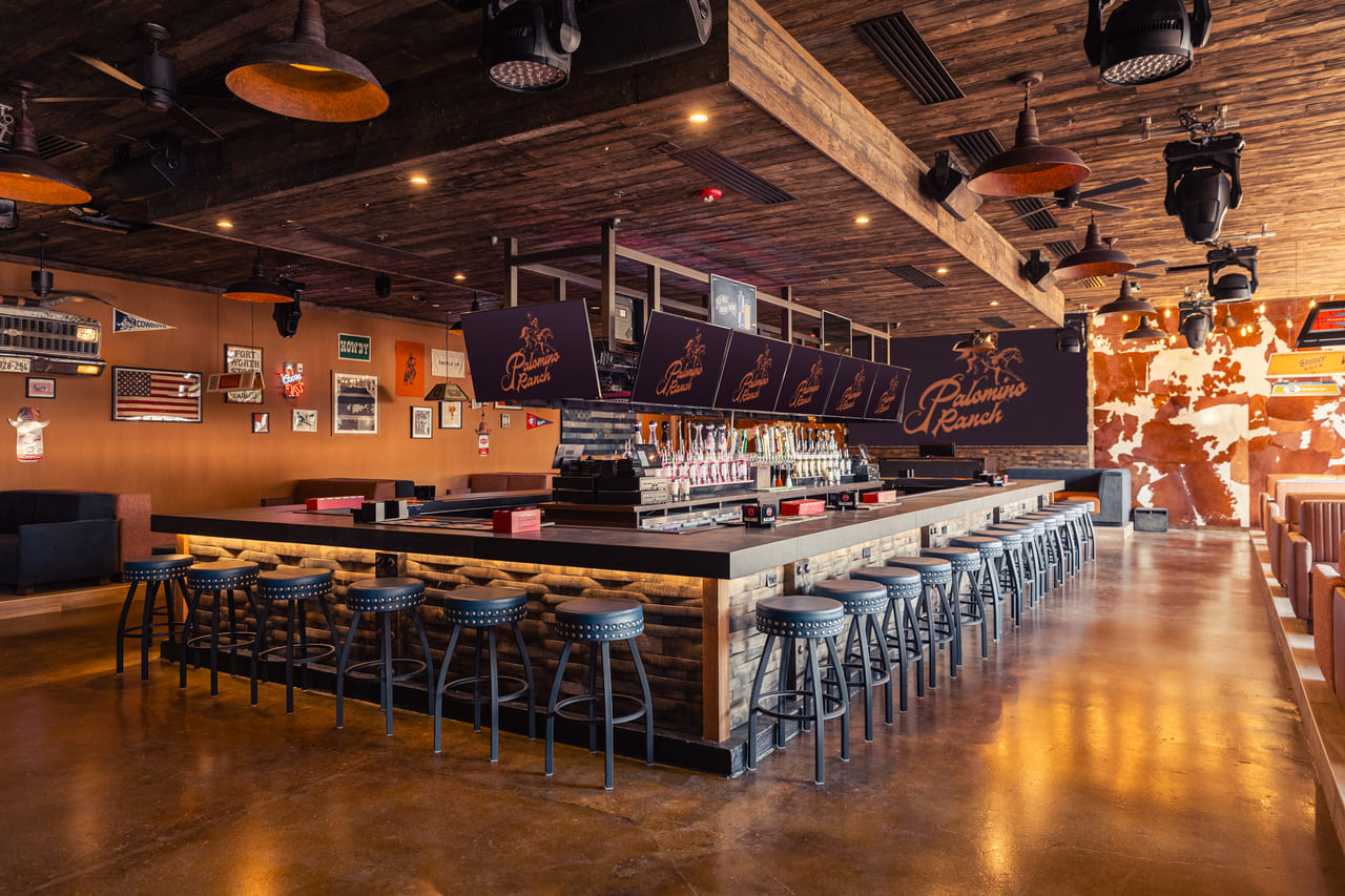 View of a rustic indoor bar with cowhide wall decor, string lights, brown leather booths, and a long bar with high-top stools at Palomino Ranch.