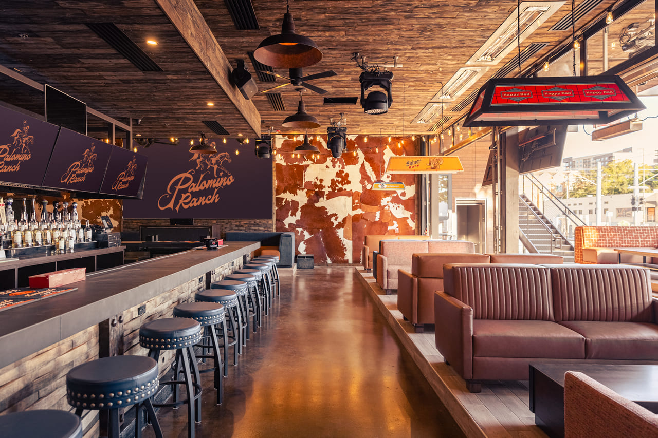 Bar view at Palomino Ranch featuring mounted TVs with logo, open layout with floor-to-ceiling windows, and cozy booth seating throughout.