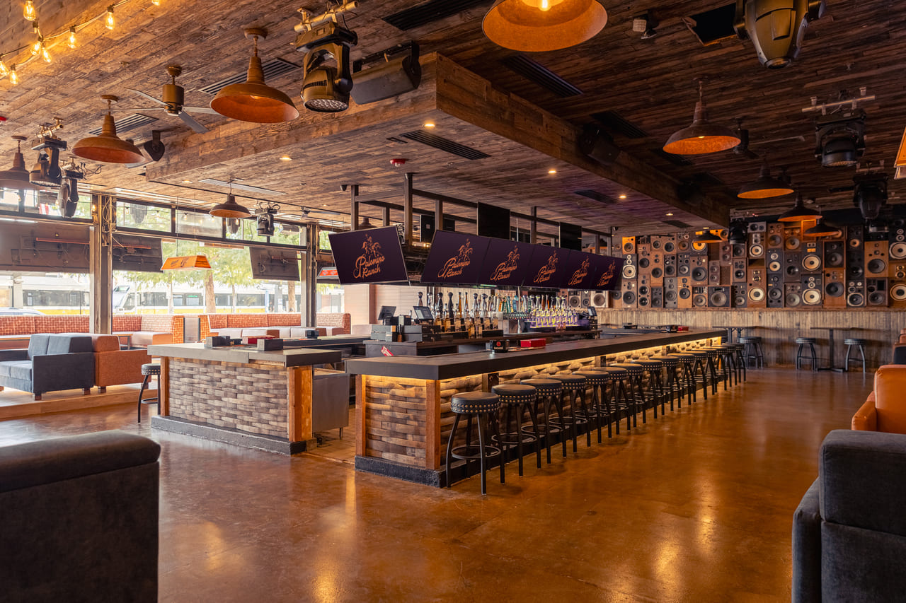 Wide-angle view of a Western-themed bar interior featuring wood-paneled ceiling, exposed beams, barstools, and vintage signage along the walls.