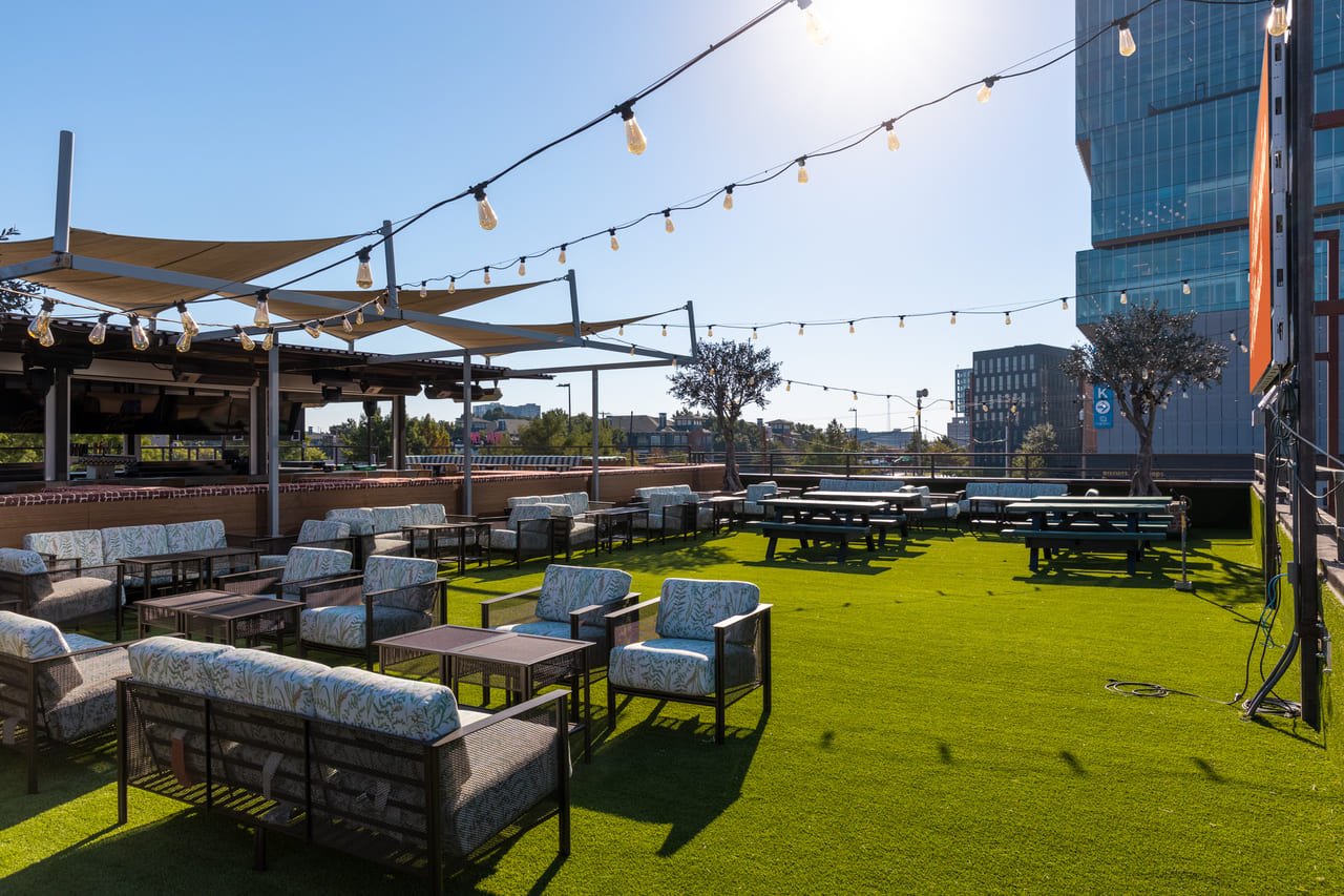 Rooftop lounge area with artificial turf, cushioned seating, string lights, and shaded canopies under a sunny sky.