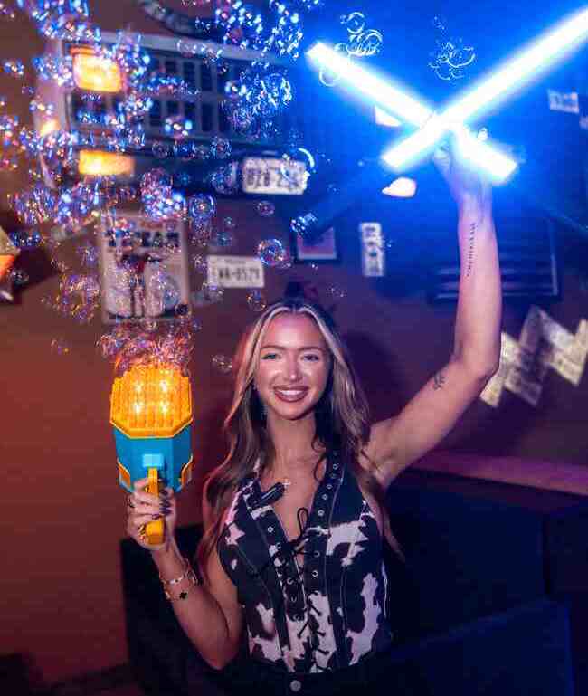 Smiling woman at a bar holding a bubble gun and two glowing blue light wands with bubbles floating in the air.