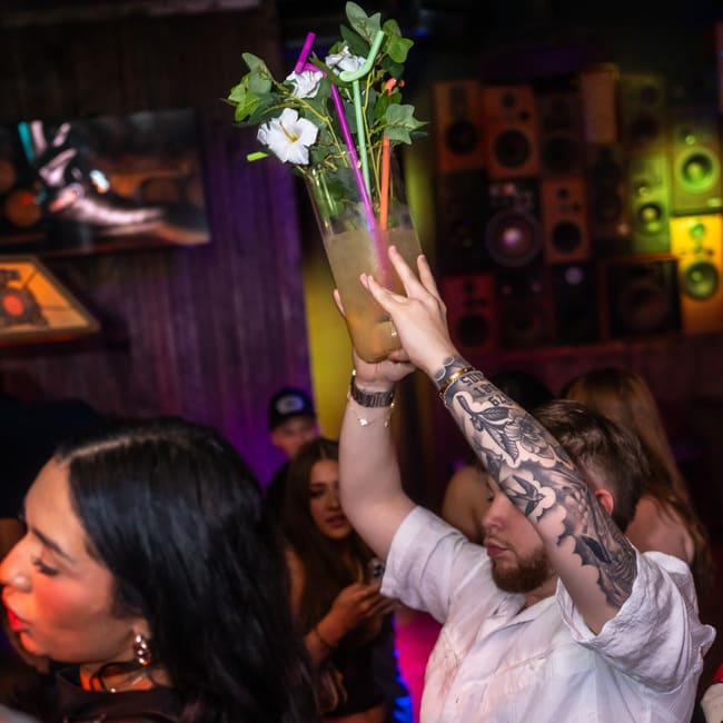 Guest holding a large cocktail pitcher with straws and herbs above the crowd in a lively nightclub.