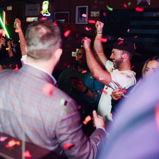 Guests celebrating in a bar as colorful confetti falls and people raise their arms in excitement.