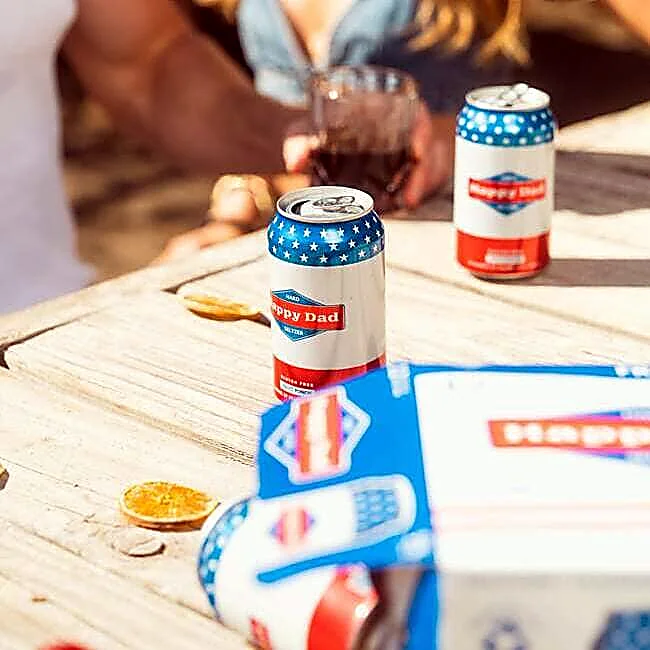Close-up of canned beverages on a wooden outdoor table during a social gathering, with guests and drinks softly blurred in the background.