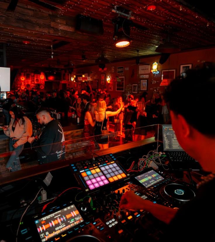DJ performing from a booth overlooking a crowded dance floor filled with guests enjoying Latin Night under warm red lighting in a lively nightclub.
